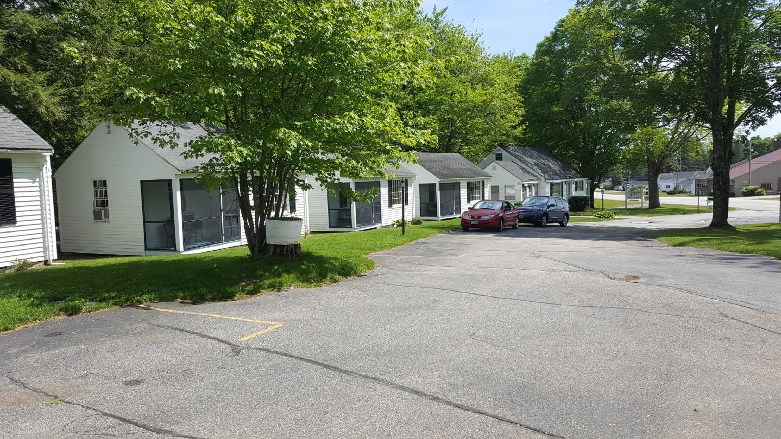 A row of single-story mobile homes with attached porches, a paved driveway, and a small parking area with two cars.