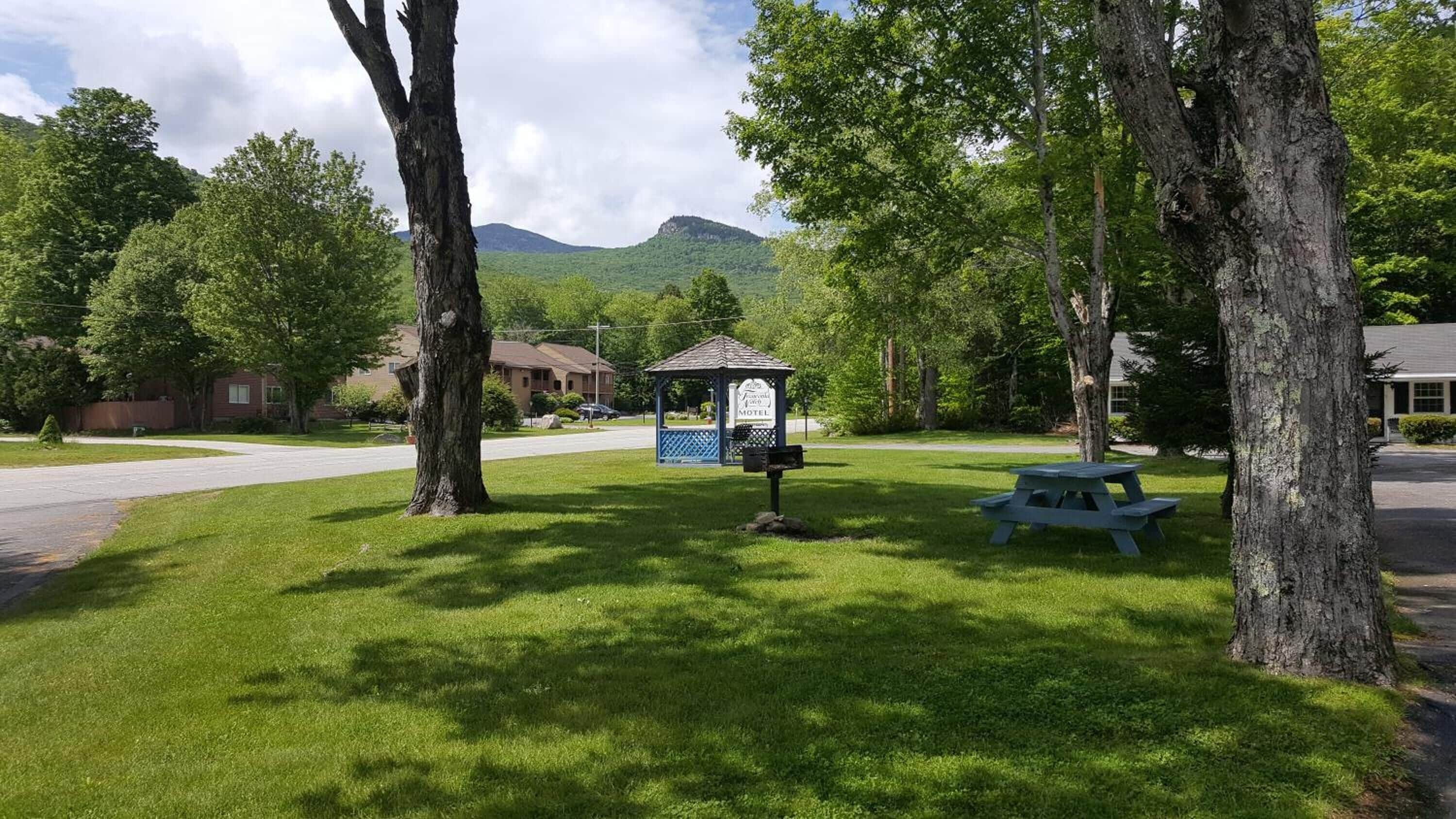 A gazebo-style structure with a sign indicating a motel, surrounded by lush green grass and trees, with a mountain in the background.