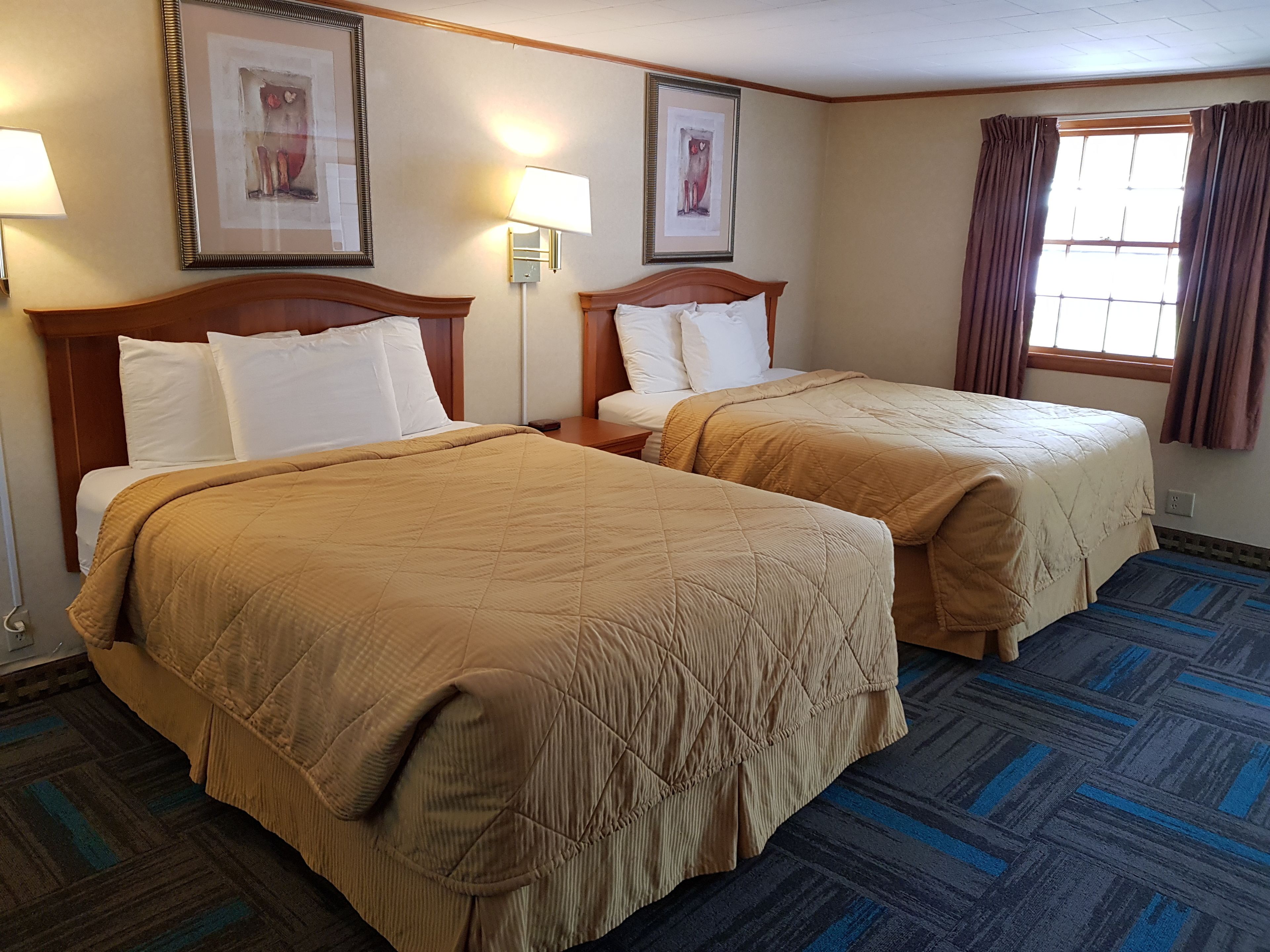 A hotel room with two beds, each with a wooden headboard, white bedding, and a brown quilt. There are framed pictures on the walls, a window with curtains, and a patterned carpet.