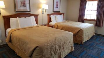 A hotel room with two beds, each with a wooden headboard, white bedding, and a brown quilt. There are framed pictures on the walls, a window with curtains, and a patterned carpet.