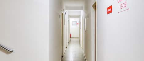 A narrow, well-lit hallway with a tiled floor, leading to a door at the end. There are signs on the wall indicating room numbers and a fire extinguisher mounted on the wall.