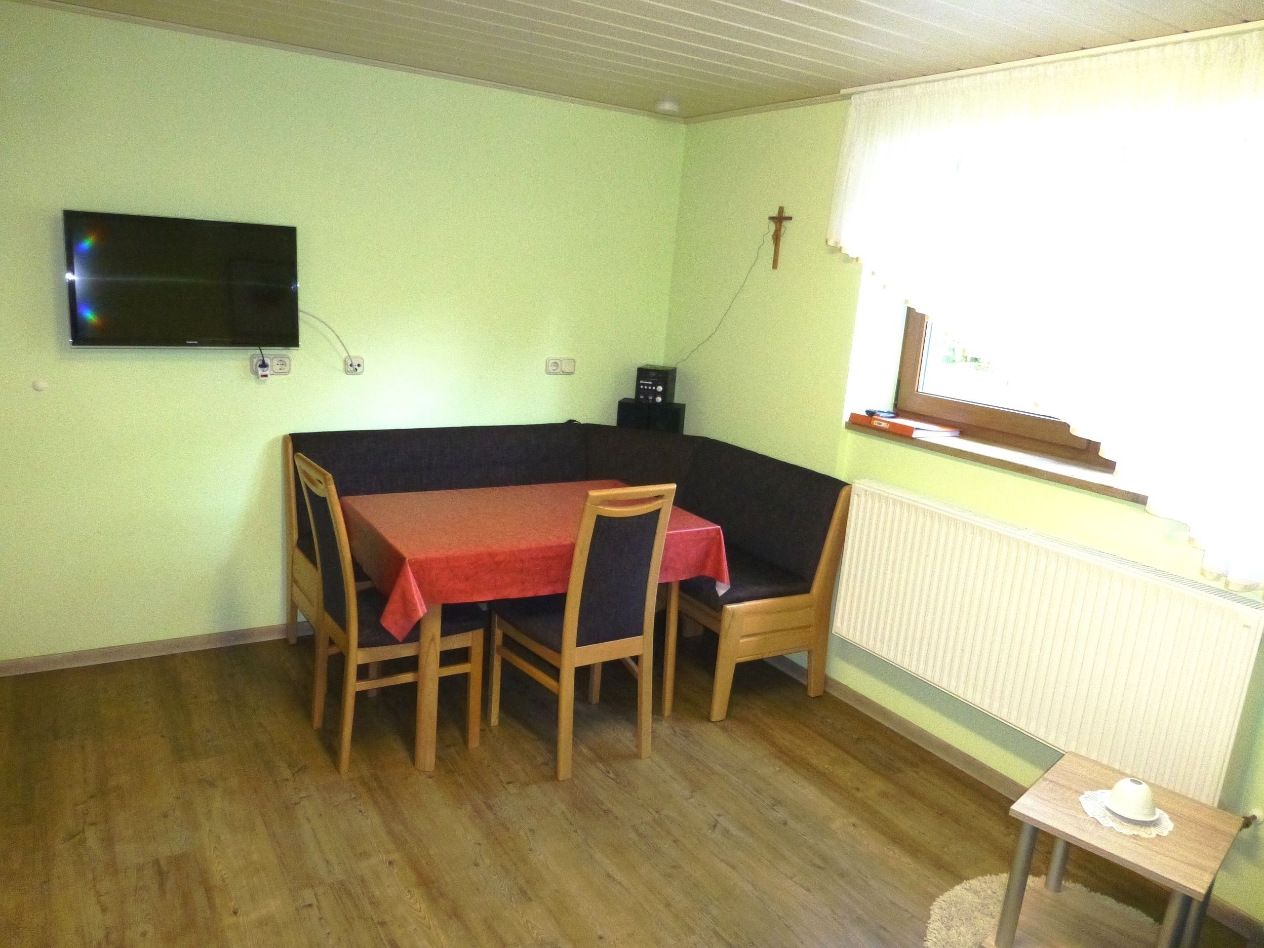 A dining area with a table and chairs, a television mounted on the wall, a cross on the wall, and a window with a view.