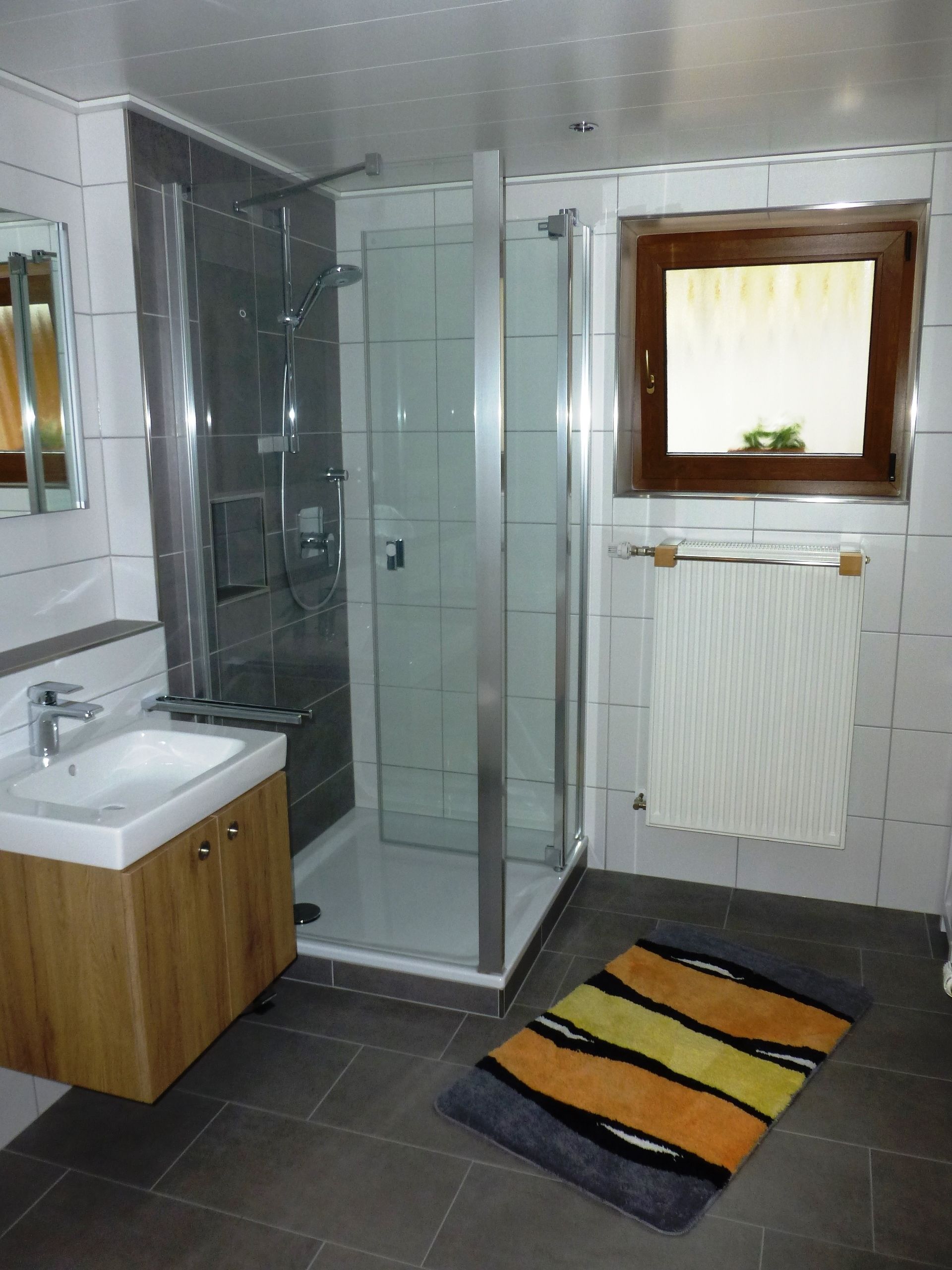 A modern bathroom with a glass shower enclosure, a sink with a wooden cabinet, and a colorful rug on the floor.