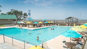 A swimming pool with multiple lanes, surrounded by umbrellas and lounge chairs, with a view of the ocean and a lifeguard tower in the background. - Sound Waves - Colington Harbor, Waterfront, Pets OK, Community Pool & Soundfront Beach (Kill Devil Hills)