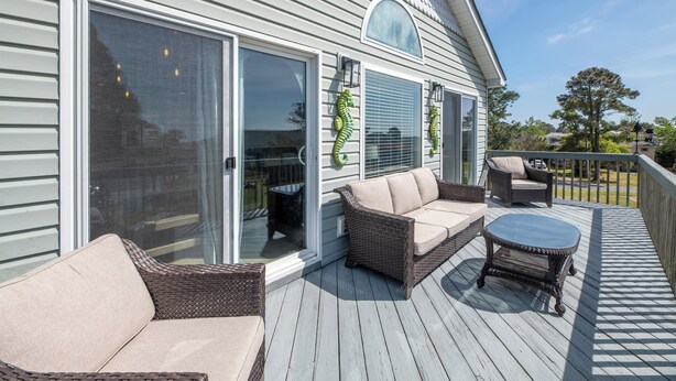 A covered deck with wicker furniture, large sliding glass doors, and a view of the surrounding landscape. - Sound Waves - Colington Harbor, Waterfront, Pets OK, Community Pool & Soundfront Beach (Kill Devil Hills)
