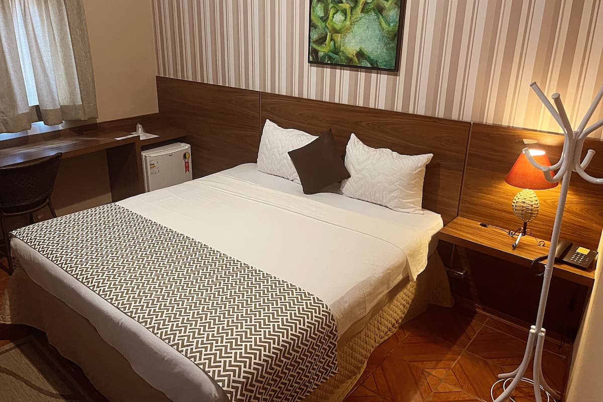 Quarto de hotel com cama, escrivaninha, cadeira, mesa de cabeceira com abajur e obra de arte na parede.