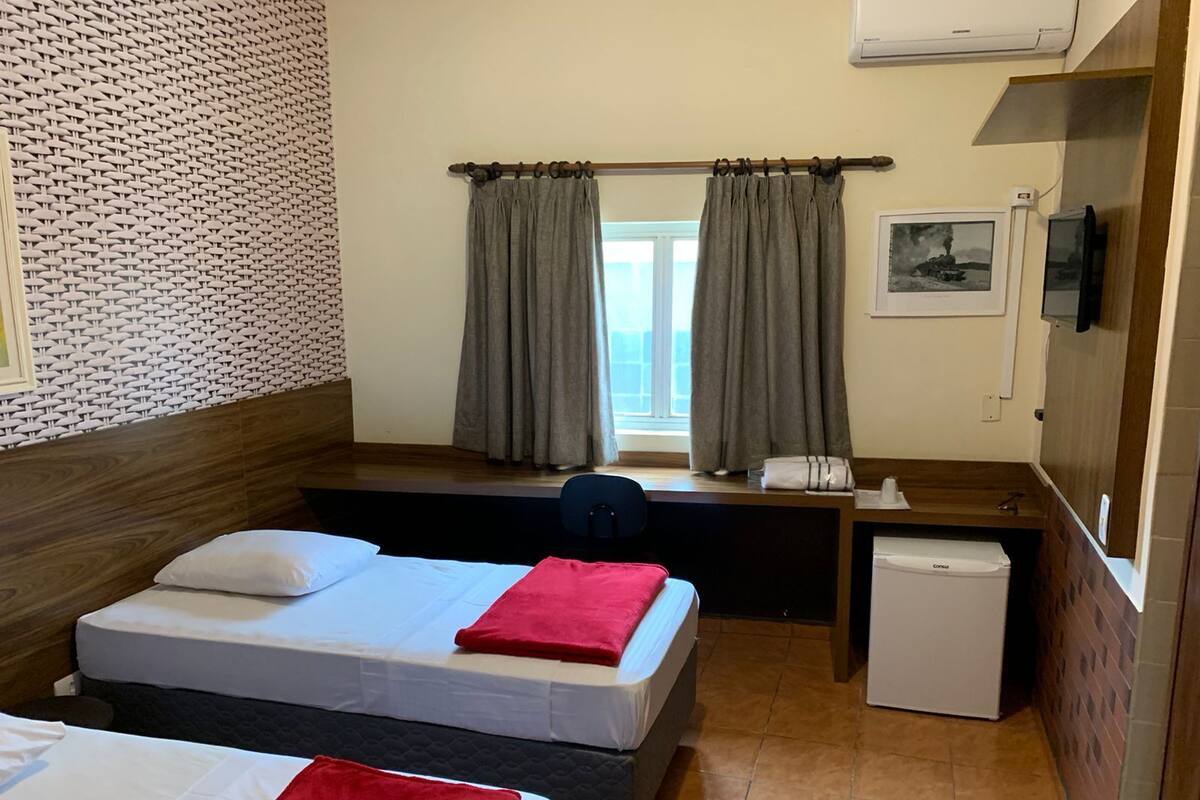 Quarto de hotel com duas camas, uma mesa pequena, um frigorífico e uma janela com cortinas.
