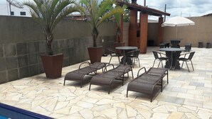 Une terrasse extérieure avec un sol en pierre, des chaises longues, une piscine et un parasol blanc.