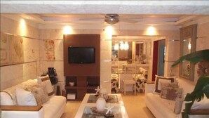 Una sala de estar con un sofá blanco, una mesa de centro con objetos decorativos, un televisor de pantalla plana montado en la pared y un ventilador de techo.