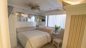 Un dormitorio con una cama grande, un ventilador de techo, una mesita con una silla y una ventana con cortinas.