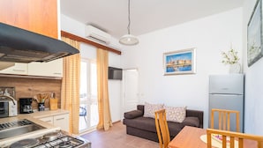 A compact kitchen with a gas stove, a sink, and a modern range hood. There is a dining area with a wooden table and chairs, and a sofa with a patterned cushion. A refrigerator is visible in the background. - Apartments Senjo - One Bedroom Apartment with Balcony and Sea View (4 adults) (Cavtat)