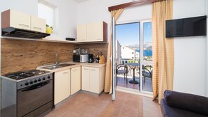 A compact kitchen with a stove, sink, and cabinets, leading to a balcony with a table and chairs. - Apartments Senjo - One Bedroom Apartment with Balcony and Sea View (4 adults) (Cavtat)