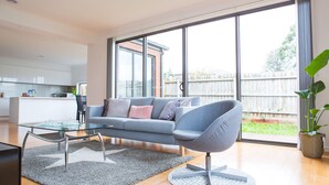 A modern living room with a glass coffee table, grey sofa, and a large window with a view of a backyard. - 5 Bedroom | 3 Bathroom Holiday Home near Glen Waverley with WIFI and NETFLIX (Forest Hill)