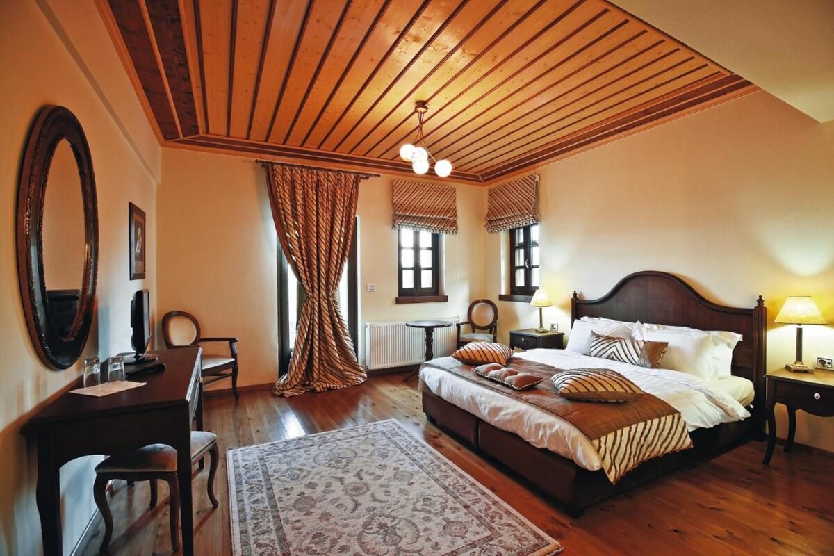 A bedroom with a wooden ceiling, a large bed with multiple pillows, a desk with a chair, and a large mirror.
