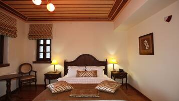 Un dormitorio ordenado con techo de madera, una cama grande con almohadillas a rayas, dos mesitas de noche con lámparas, una silla y una ventana con persianas.