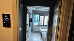 A hotel hallway leading to a bedroom with a bed, a window with blinds, and a door with a number plate. - Grand FM Hotel (Istanbul)