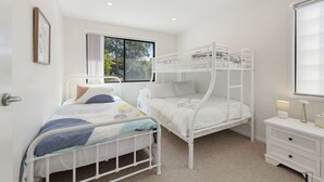 A bedroom with a bunk bed, a single bed, a bedside table, and a window with a view of trees. - Seaside Living, Steps to the Beach (LONG BEACH)