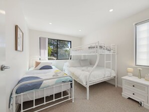 A bedroom with a bunk bed, a single bed, a bedside table, and a window with a view of trees. - Seaside Living, Steps to the Beach (LONG BEACH)