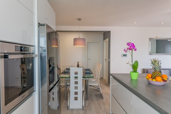 A modern kitchen with stainless steel appliances, a dining area with a glass table, and a fruit bowl on the counter.