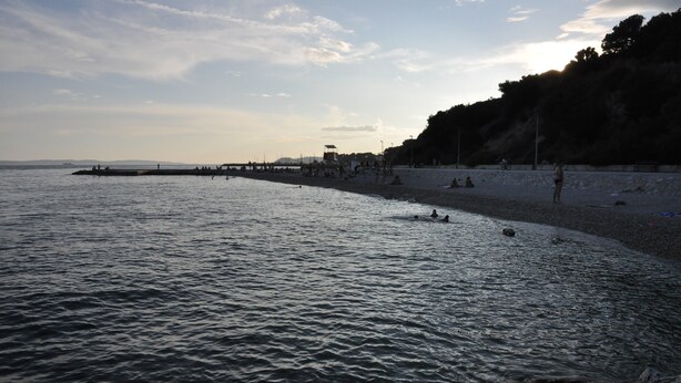 Una playa con gente nadando y relajándose en la arena, un muelle que se adentra en el agua y un acantilado rocoso al fondo.