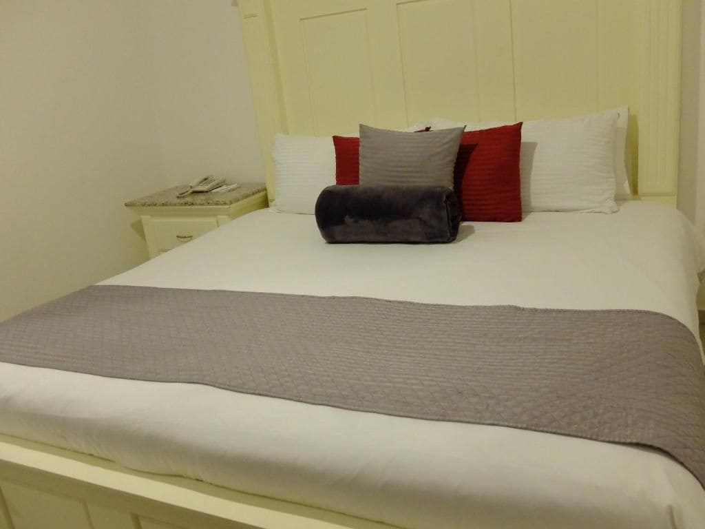 Una cama con ropa blanca, un edredón gris y varias almohadas rojas y grises.