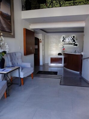 Un vestíbulo de hotel con una mesa de recepción, un área de descanso con un sofá y una mesa, y una escalera que conduce a un nivel superior.