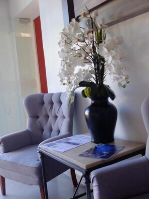 Un espace d’attente comprenant un fauteuil capitonné, une petite table ornée d’un vase de fleurs blanches et un tableau encadré posé sur la table.