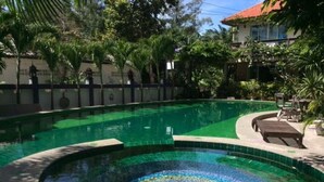 Piscina com design em mosaico ao centro, rodeada por palmeiras e um edifício com telhado vermelho.