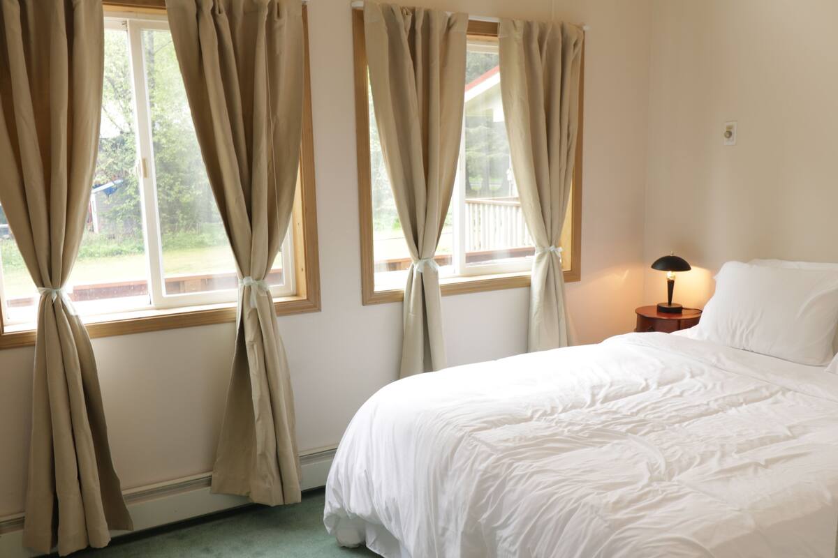 A bedroom with a bed, curtains tied at the windows, a bedside lamp, and a view of the outdoors.