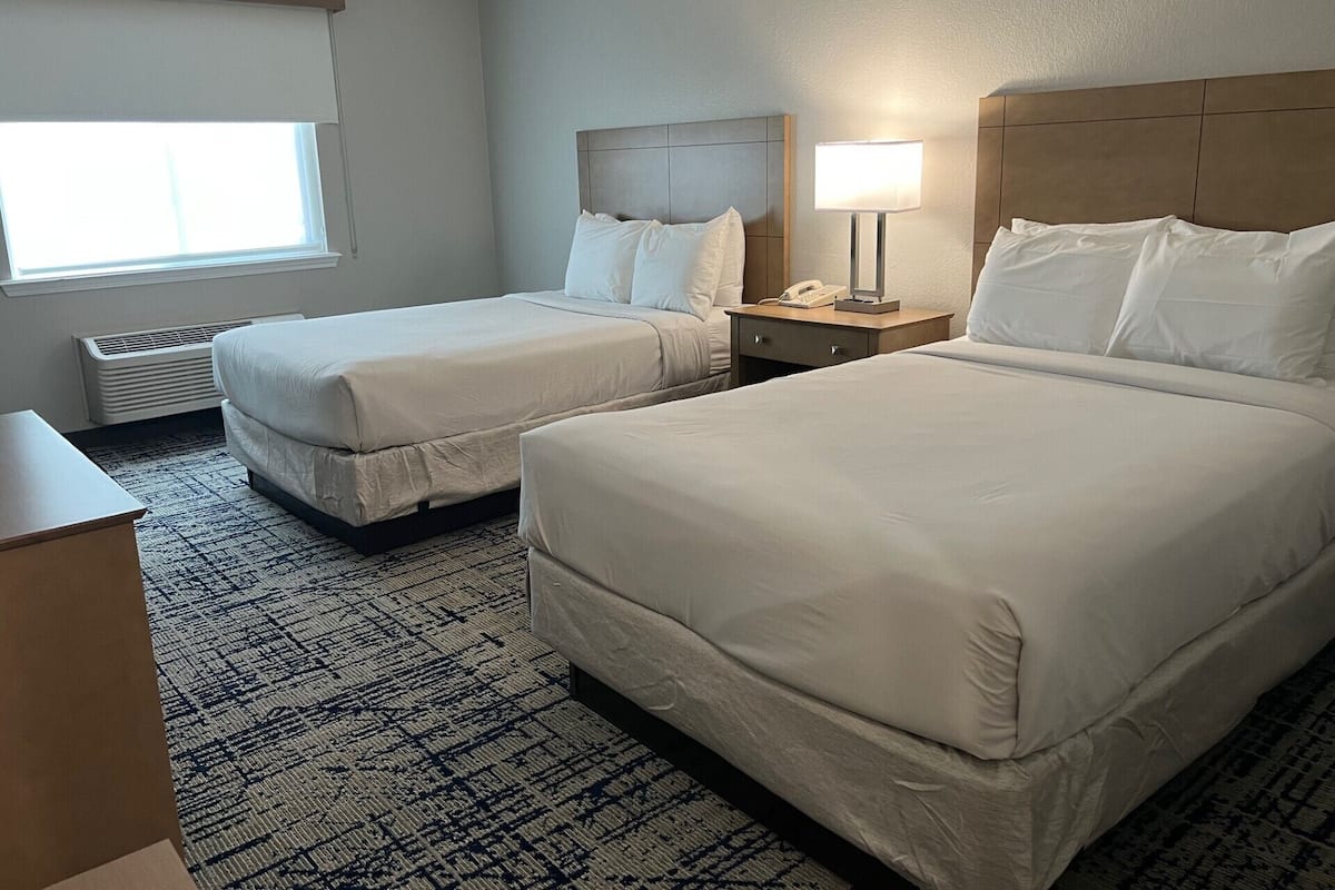 Quarto de hotel com duas camas, uma mesa de cabeceira com abajur, telefone e janela com persianas.