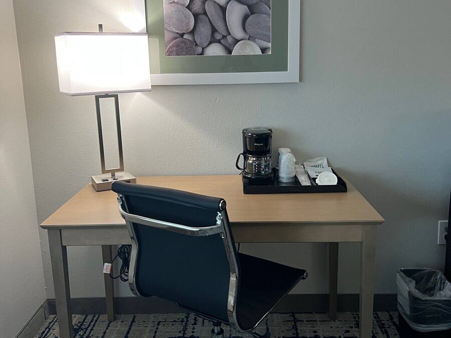 A hotel room with a desk, a chair, a lamp, a coffee maker, and a framed picture on the wall.