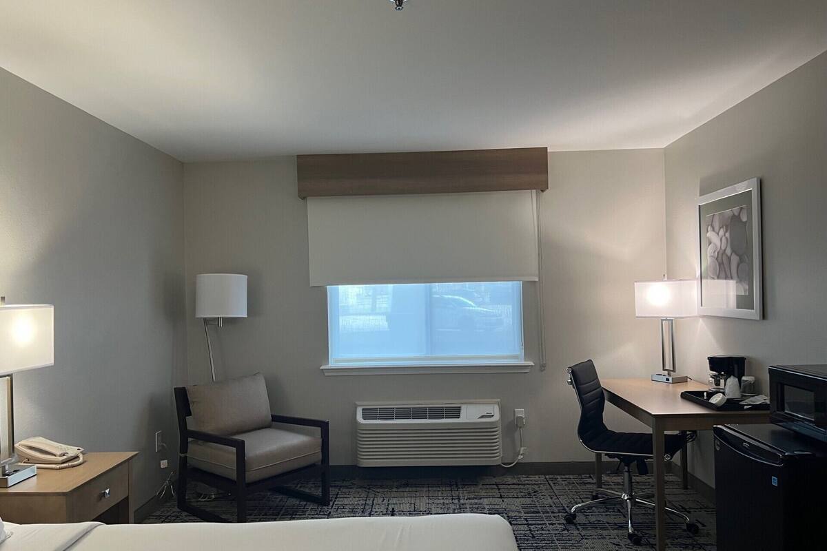 Quarto de hotel com cama, escrivaninha, cadeira, abajur, aquecedor e janela com persianas.