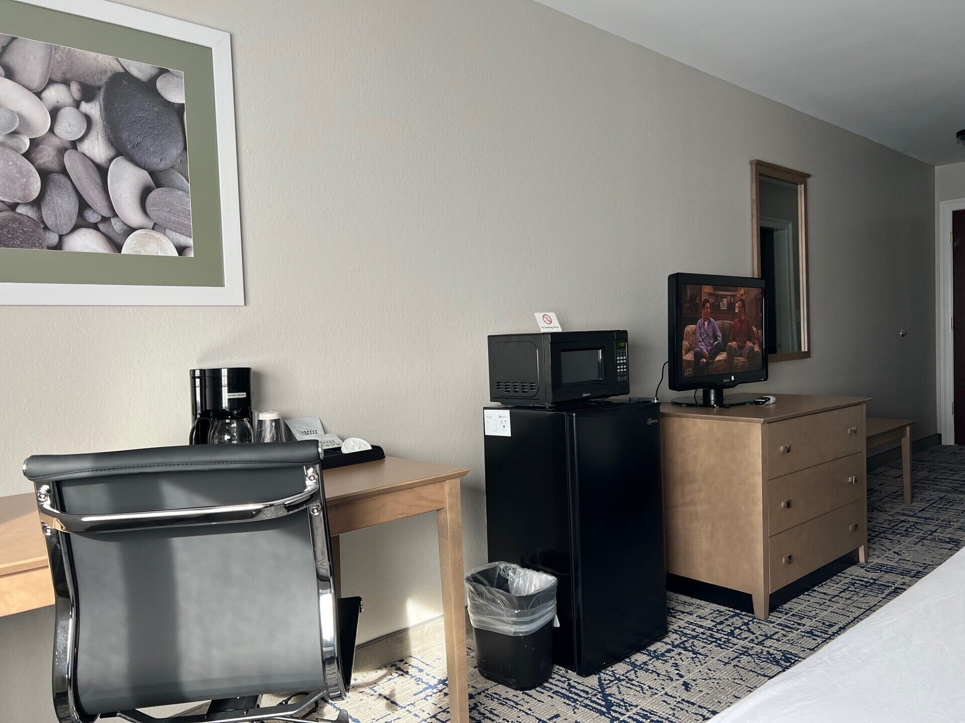Een hotelkamer met een bureau, een zwarte koelkast, een magnetron, een televisie, een prullenbak en een bed.