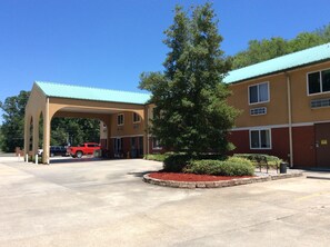A building with a covered parking area, a large tree, and a red vehicle in the background.