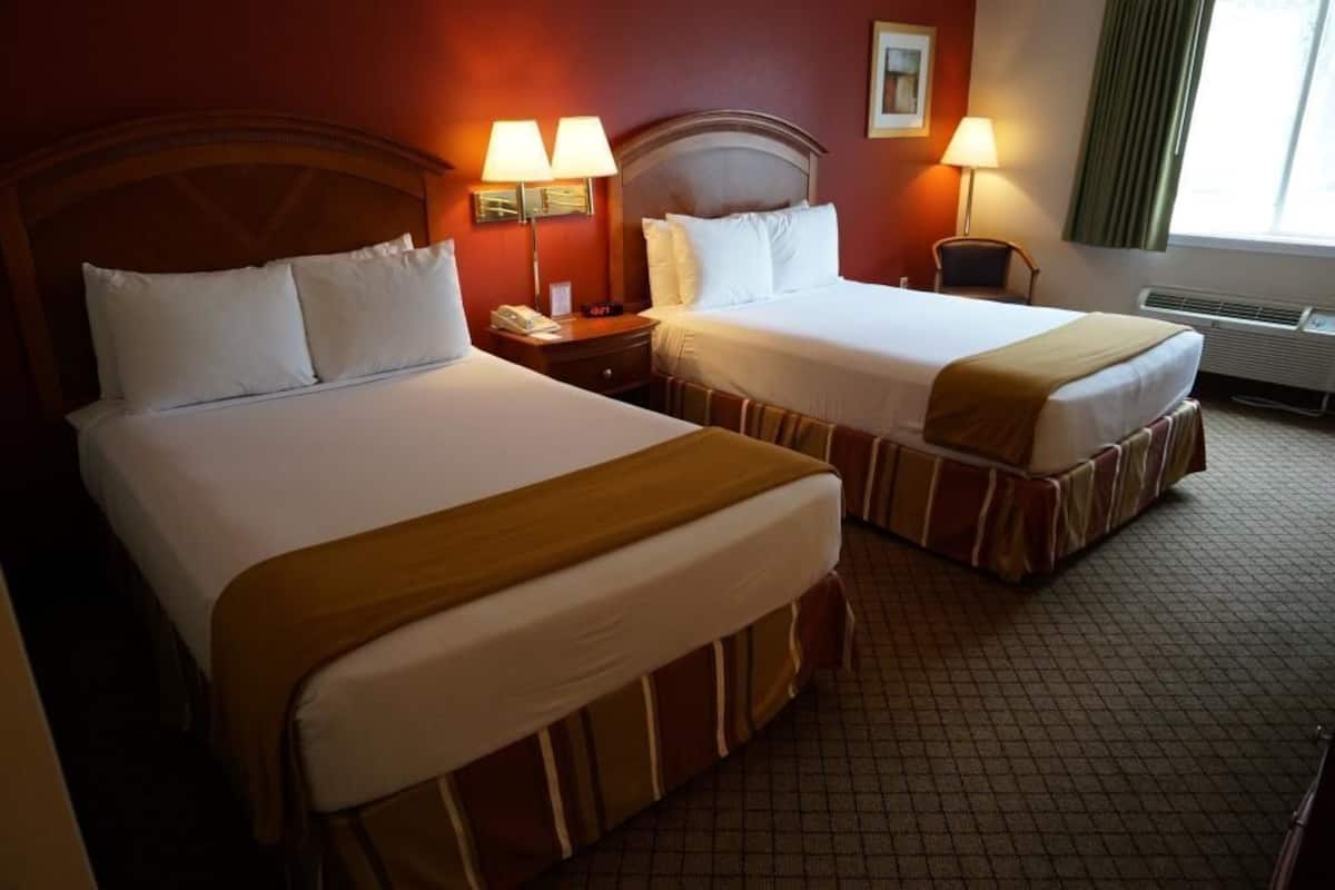 Quarto de hotel com duas camas, uma mesa de cabeceira com abajur, uma cadeira e uma janela com cortinas.