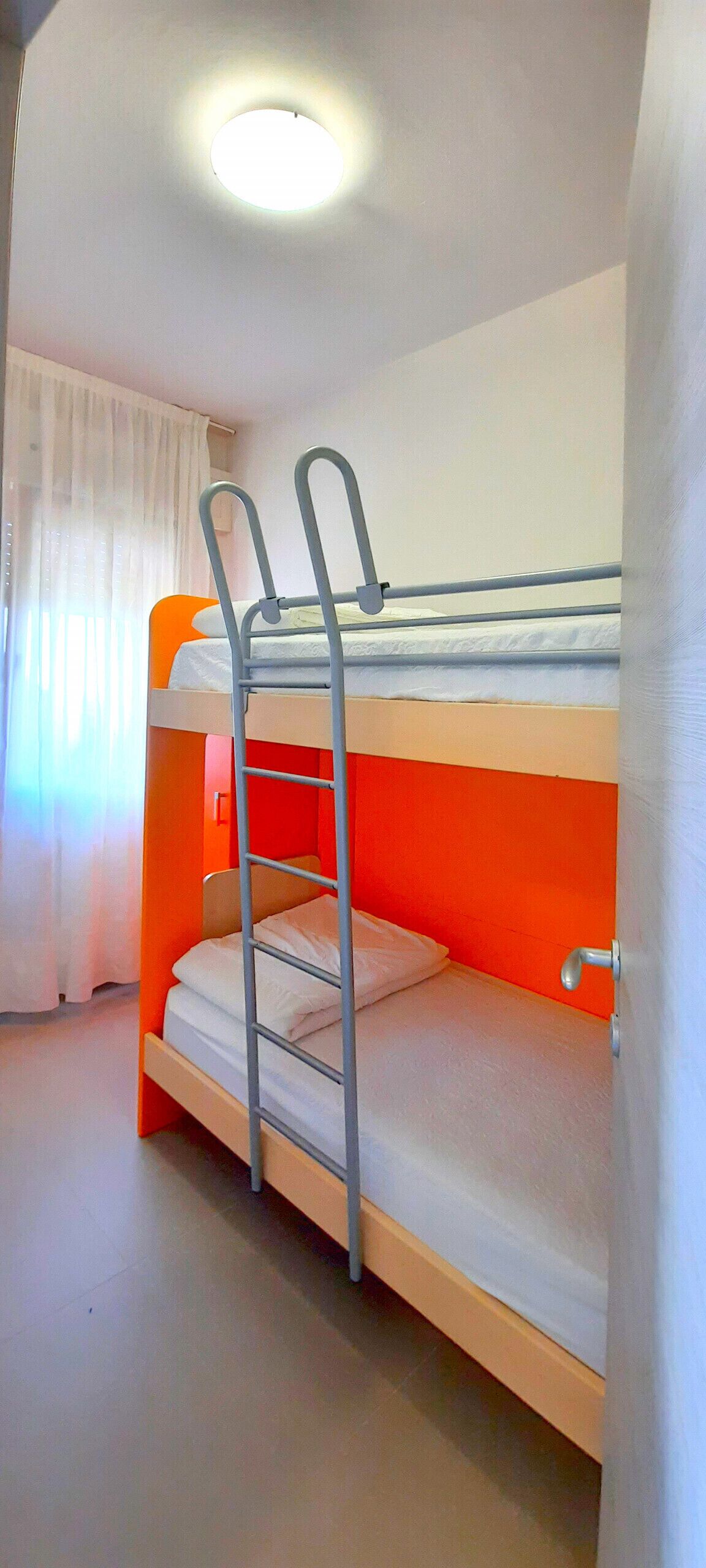 Uma cama beliche com escada, uma janela com cortinas e uma luminária no teto.