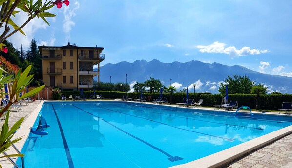 A swimming pool with clear blue water, surrounded by lounge chairs and a building with a mountain view in the background.