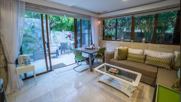 Un salón amplio con sofá, mesa de centro y comedor con sillas y mesa. Ventanas grandes ofrecen vistas al exterior.