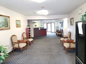 A hotel lobby with a reception desk, several wooden chairs, a patterned carpet, and framed artwork on the walls.