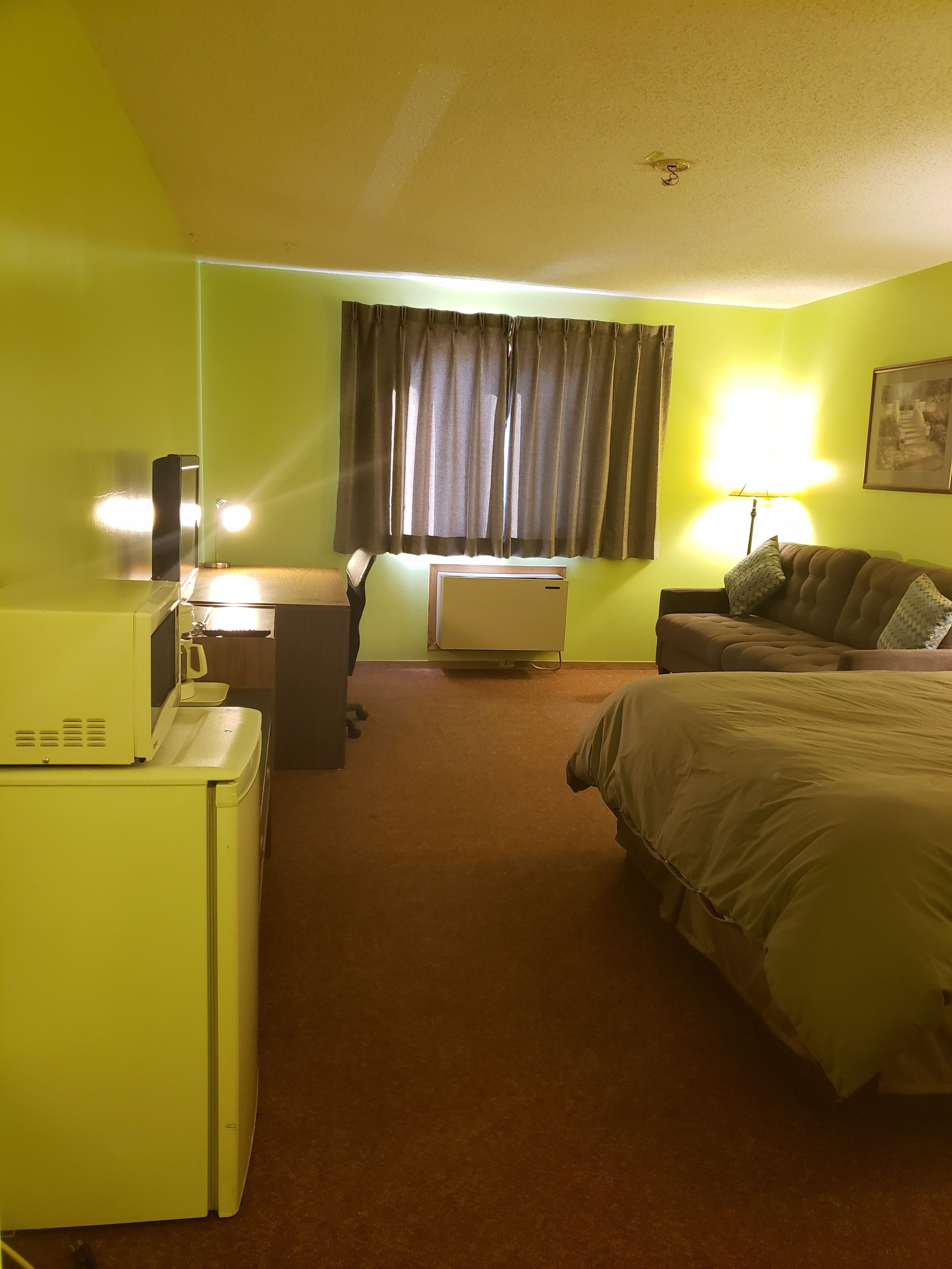 Ett hotellrum med en säng, ett skrivbord med en lampa, en mikrovågsugn och ett fönster med gardiner.