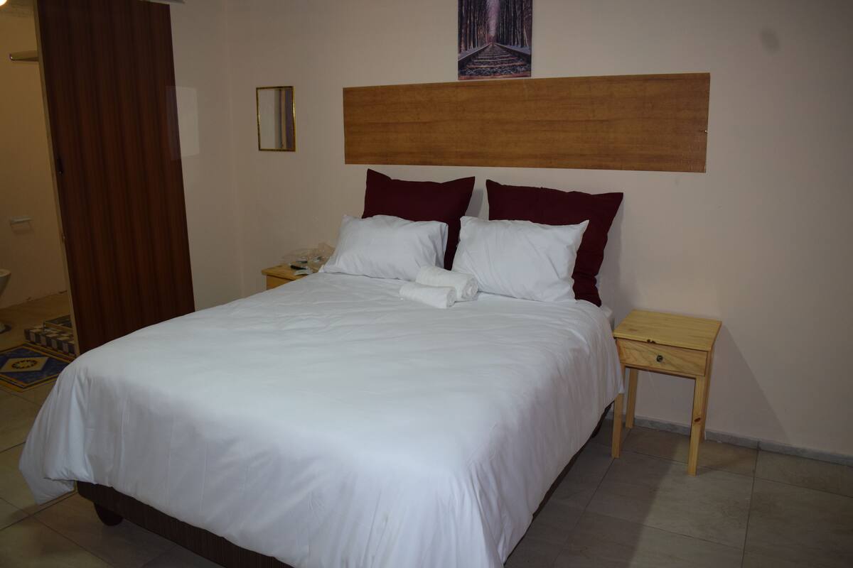 A neatly made bed with white and maroon bedding, a wooden headboard, and a small wooden nightstand beside it.