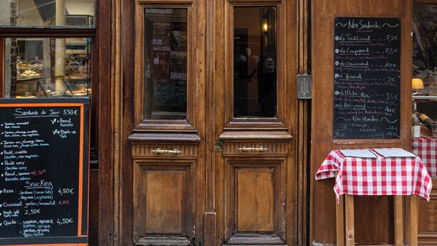 A wooden storefront with a chalkboard menu, a small table with a checkered tablecloth, and a red and white striped awning.