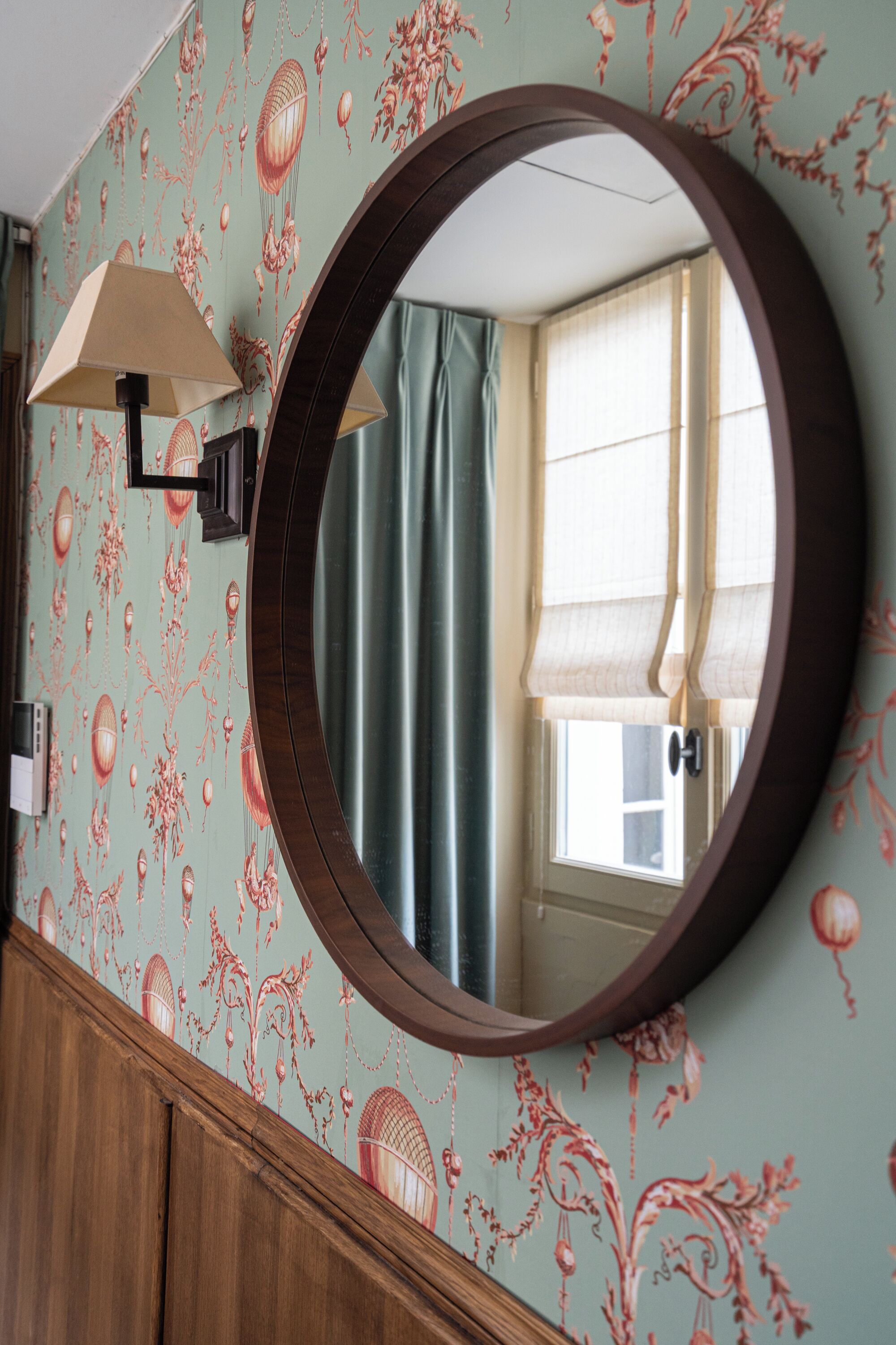 Ein Badezimmer mit rundem Spiegel, floralem Wandpapier, einem Holzwaschtisch und einem Fenster mit Vorhängen.