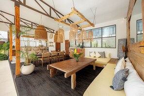 A spacious room with a wooden dining table, a sofa with patterned cushions, and a large window providing natural light.