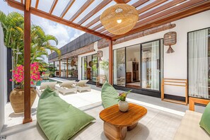 An outdoor seating area with a wooden table and chairs, a large vase with flowers, and a pergola with a woven ceiling.