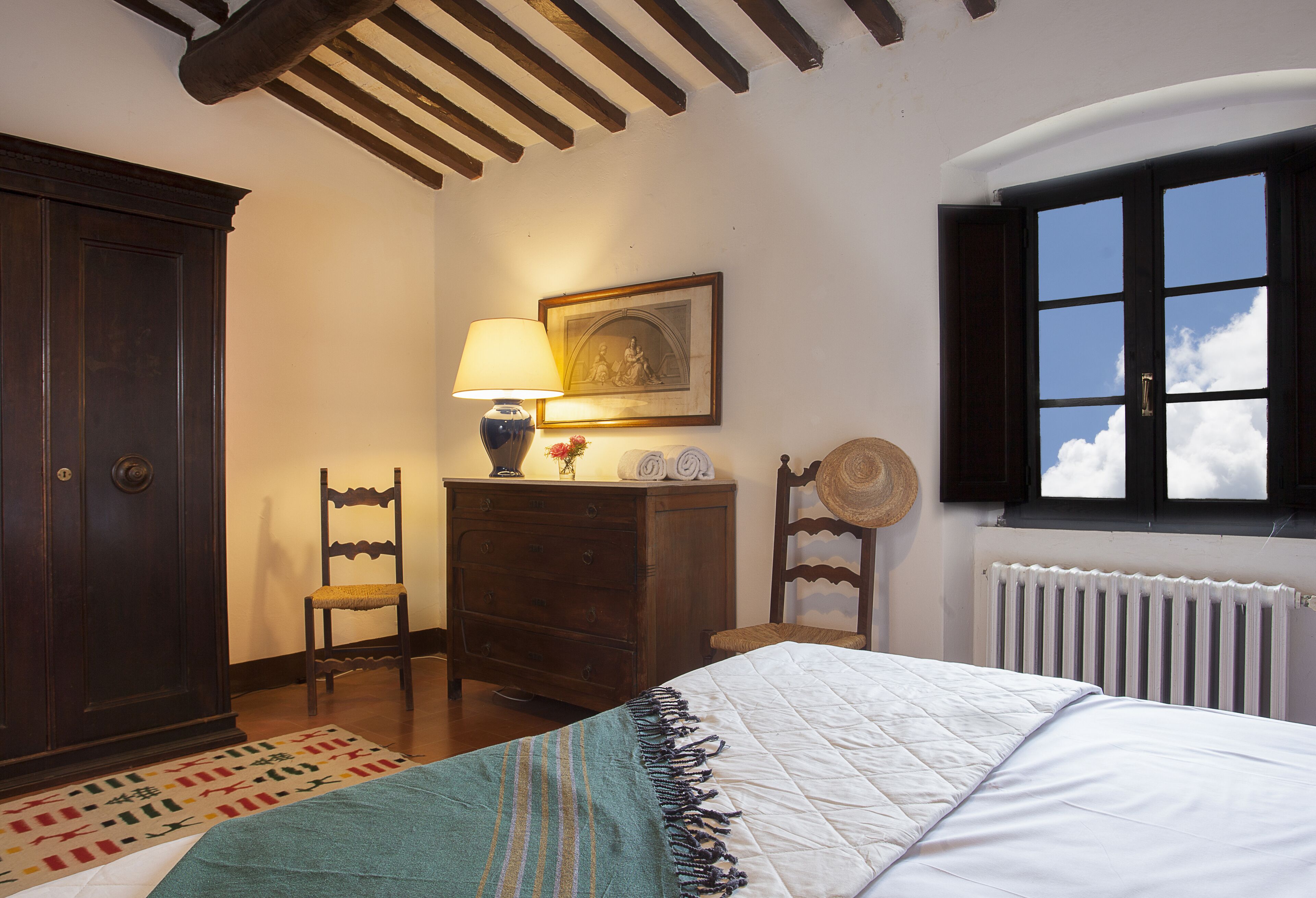 A bedroom with a bed, a dresser, a chair, a lamp, a picture, and a window with a view of the sky.