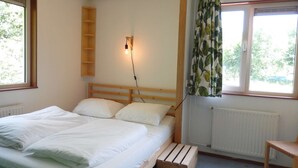 A bedroom with a bed, a wooden headboard, a nightstand, a wall-mounted light, a window with curtains, and a radiator. - Amsterdam Farm Lodge: cosy holliday home (Amsterdam)