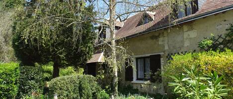 Une maison à deux étages, dotée d’un toit en pente, entourée d’une végétation luxuriante et d’arbres.