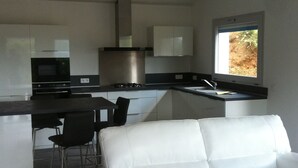 Cocina moderna con horno empotrado, estufa a gas y campana extractora de acero inoxidable. Cuenta con un comedor con sofá blanco y dos sillas negras.