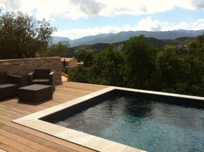Une piscine dotée d’une terrasse en bois et de meubles en osier, entourée de verdure et avec des montagnes en toile de fond.
